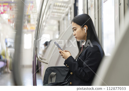 Image of a high school girl wearing earphones looking at the screen of a smartphone on the train commuting to school by train 112877033