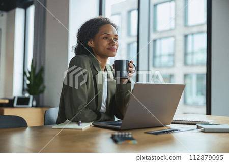 Woman seated at a table with a laptop and a cup of coffee 112879095
