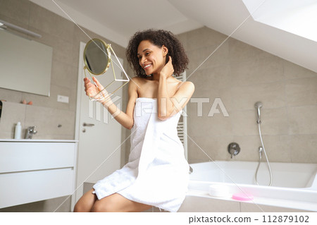 A woman, wrapped in a towel, sits in front of a mirror in the bathroom 112879102