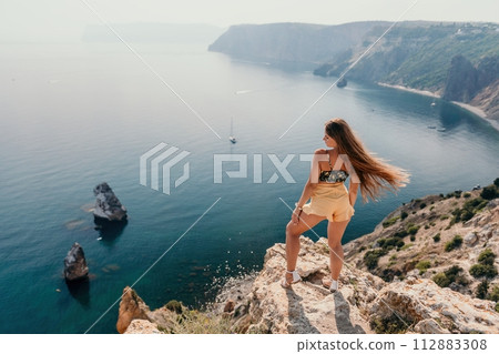 Woman travel sea. Happy tourist enjoy taking picture outdoors for memories. Woman traveler looks at the edge of the cliff on the sea bay of mountains, sharing travel adventure journey 112883308