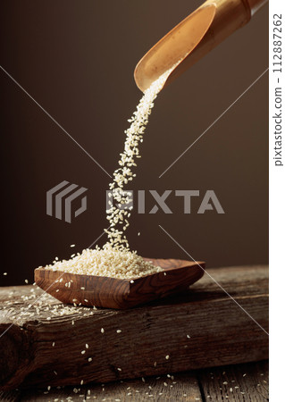 Grains of white sesame are poured with a wooden spoon in the wooden dish. 112887262