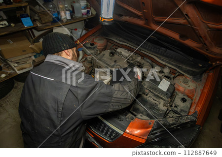 Top view of a driver pouring oil into the engine under the hood of a car in his garage. A man holds a canister of oil in his hands and carefully pours it into the engine of a car in a garage. 112887649