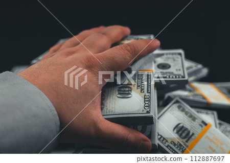 First person. A man reaches out and takes a wad of dollars from a pile on a table against a black background. Close-up. A hand reaches out and takes a wad of money from a table on a black background. 112887697