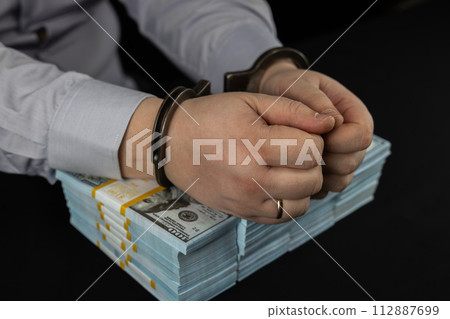A corrupt official in handcuffs put his hands on stacks of dollars on a black background. Purchase plan of handcuffed hands and a large number of dollars. A corrupt official in handcuffs put his hands on stacks of dollars on a black background. Purchase plan of handcuffed hands and a large number of dollars. 112887699