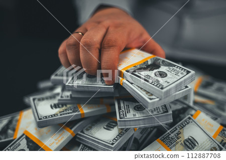 Beautiful advertisement. A man's hand takes out a wad of dollars from a pile of money, close-up. A hand takes money on a black background. The concept of big earnings, wealth, financial well-being. 112887708