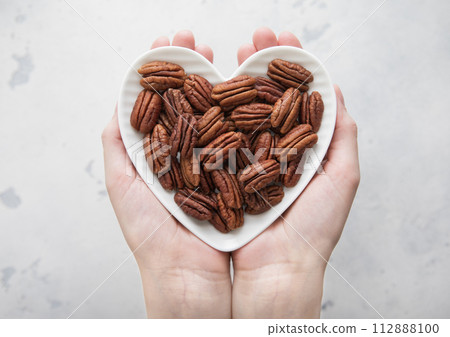 Female hands holding heart shaped plate with peeled pecan nuts on light background. 112888100