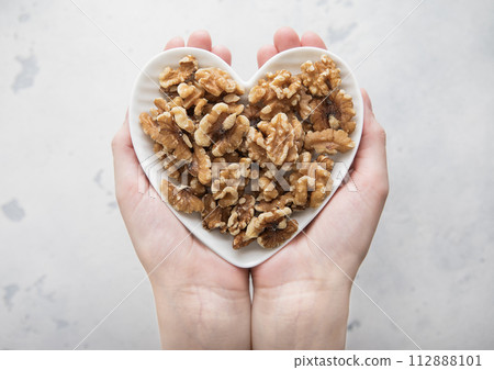 Female hands holding heart shaped plate with healthy peeled walnut nuts on light background. Female hands holding heart shaped plate with healthy peeled walnut nuts on light background. 112888101