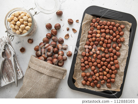 Baking tray with raw hazelnut nuts with cracker and peeled nuts in jar on light kitchen background. Baking tray with raw hazelnut nuts with cracker and peeled nuts in jar on light kitchen background. 112888103
