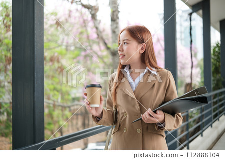 A young Asian businesswoman wearing a suit holding a cup of coffee and files walking in a big city. Young Asian businesswomen walking to the office. 112888104