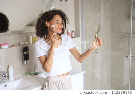 A woman smiling as she applies cream to her face in front of a mirror A woman smiling as she applies cream to her face in front of a mirror 112889176
