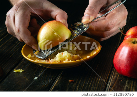 Close-up of a chef hands using a grater to grate an apple on the kitchen table to prepare a healthy meal or applesauce Close-up of a chef hands using a grater to grate an apple on the kitchen table to prepare a healthy meal or applesauce 112890780