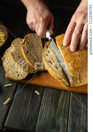Slicing bread with a knife in the cook hand on a kitchen cutting board before dinner. Low key bakery hearty food concept. Advertising space Slicing bread with a knife in the cook hand on a kitchen cutting board before dinner. Low key bakery hearty food concept. Advertising space 112890781