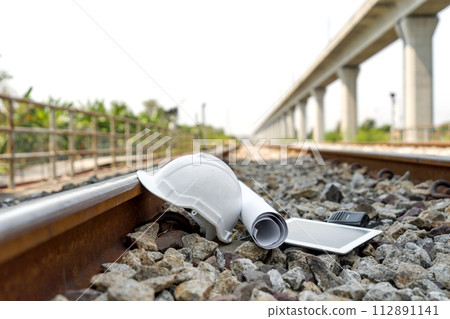 A white safety helmet, blueprint, tablet computer and a portable radio or walkie-talkie, all resting on a bed of rough stone in a railroad track. 112891141