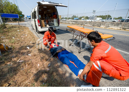 Two paramedic or emergency medical technician (EMT) in orange uniform helping neck and head accident victim lying on stretcher long spinal board. Urgent assistance during road accident. Two paramedic or emergency medical technician (EMT) in orange uniform helping neck and head accident victim lying on stretcher long spinal board. Urgent assistance during road accident. 112891322