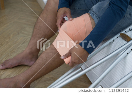 A man with a broken knee sitting on his bed. He leans on crutches, signalling recovery from injury. His bedroom serves as a backdrop, tinged with hints of helplessness yet enduring determination. A man with a broken knee sitting on his bed. He leans on crutches, signalling recovery from injury. His bedroom serves as a backdrop, tinged with hints of helplessness yet enduring determination. 112891422