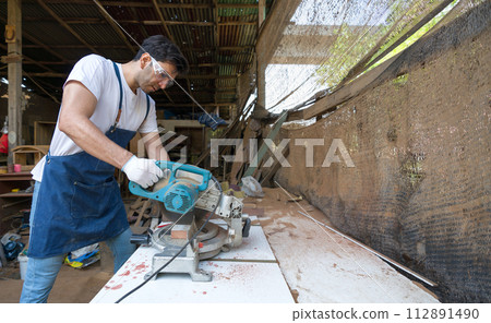 Young carpentry using a power miter saw to cut a piece of wood. Atmosphere in a workshop or outdoor shed based on the rustic environment and the presence of tools and construction materials. 112891490