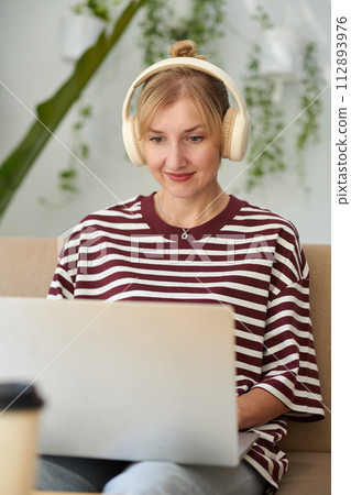 Portrait of smiling woman listening to music in headphones when coding on laptop at home 112893976