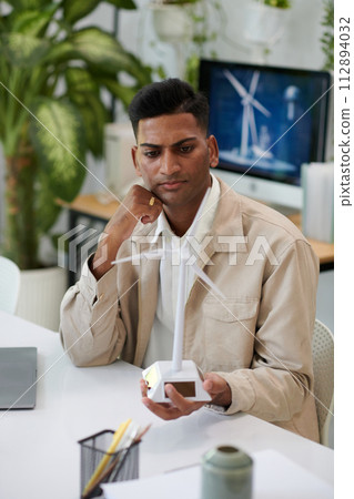 Pensive engineer looking at small plastic model of wind turbine Pensive engineer looking at small plastic model of wind turbine 112894032