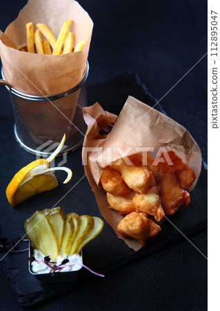 Fried chicken wings on the table. Crispy fried chicken in batter. French fries in paper. Slices of pickled cucumber. Fast food. 112895047