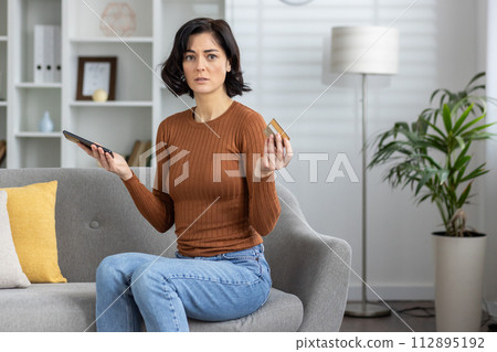 A woman holds a smartphone and credit card feeling confused and concerned about an error during an online ,transaction while sitting on the couch at home. A woman holds a smartphone and credit card feeling confused and concerned about an error during an online ,transaction while sitting on the couch at home. 112895192