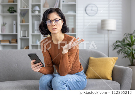 Young woman sitting on sofa, looking confused while reading a text message that seems to deliver bad news. Young woman sitting on sofa, looking confused while reading a text message that seems to deliver bad news. 112895213