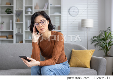 A woman sits on her couch at home, looking at her phone with a confused expression, possibly after receiving bad news. A woman sits on her couch at home, looking at her phone with a confused expression, possibly after receiving bad news. 112895214