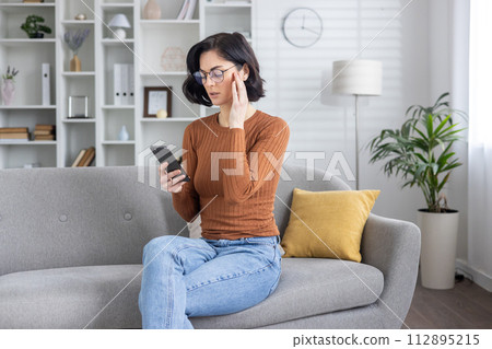 Adult female sits on the sofa at home, reading a message on her smartphone, appearing confused and concerned after receiving bad news. Adult female sits on the sofa at home, reading a message on her smartphone, appearing confused and concerned after receiving bad news. 112895215
