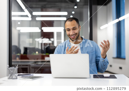 Cheerful male professional engaging in a video conference on a laptop in a well-lit, contemporary workspace. 112895279