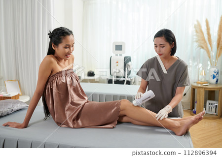 Smiling woman looking at beautician waxing her legs Smiling woman looking at beautician waxing her legs 112896394