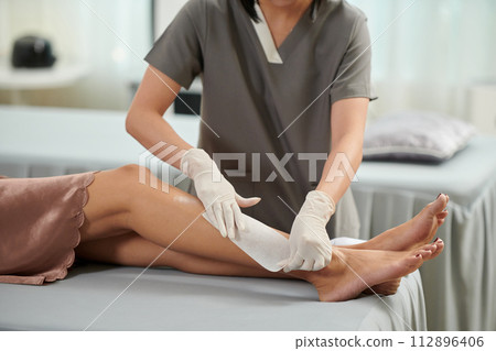 Beautician removing hair from legs of female client with wax stripe Beautician removing hair from legs of female client with wax stripe 112896406