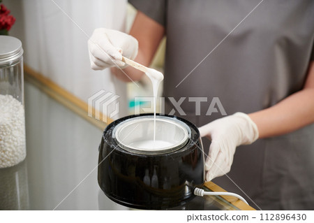 Beautician wearing gloves when mixing melted wax before applying on skin of client 112896430