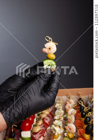 Canape, hands in black gloves hold a beautiful canape on a cap. Catering, snacking, food. Studio photo. Canape, hands in black gloves hold a beautiful canape on a cap. Catering, snacking, food. Studio photo. 112897518