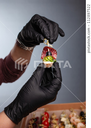 Canape, hands in black gloves hold a beautiful canape on a cap. Catering, snacking, food. Studio photo. Canape, hands in black gloves hold a beautiful canape on a cap. Catering, snacking, food. Studio photo. 112897522