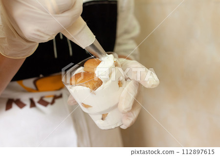 The pastry chef decorates the tiramisu in a glass with whipped cream. The pastry chef decorates the tiramisu in a glass with whipped cream. 112897615