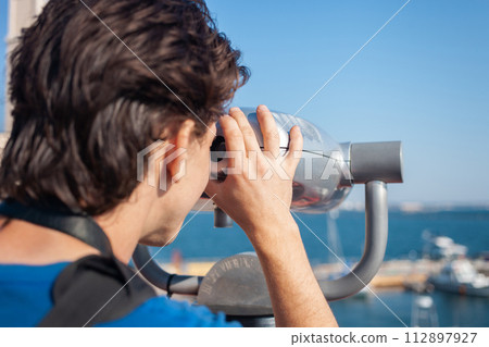 man looks through stationary binoculars. Binoscope on observation deck near sea. seaport in Odessa. Expensive yachts on the pier. Commercial machine, vending business. 112897927