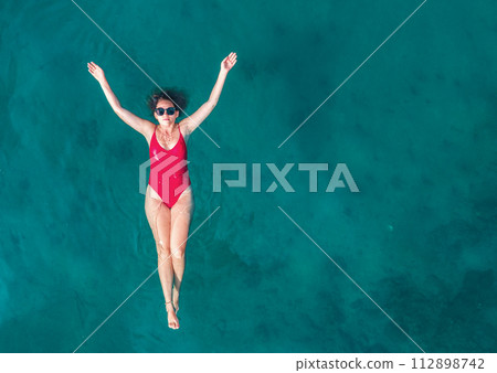 Aerial View of a Woman in Red Swimsuit Floating Serenely on the Crystal Clear Ocean Waters During Midday Aerial View of a Woman in Red Swimsuit Floating Serenely on the Crystal Clear Ocean Waters During Midday 112898742