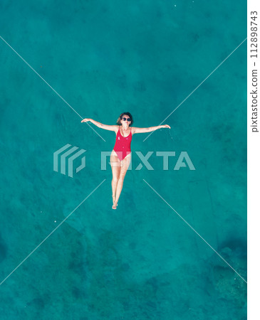 Aerial View of a Woman in Red Swimsuit Floating Serenely on the Crystal Clear Ocean Waters During Midday Aerial View of a Woman in Red Swimsuit Floating Serenely on the Crystal Clear Ocean Waters During Midday 112898743
