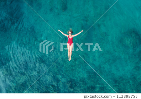 Aerial View of a Woman in Red Swimsuit Floating Serenely on the Crystal Clear Ocean Waters During Midday Aerial View of a Woman in Red Swimsuit Floating Serenely on the Crystal Clear Ocean Waters During Midday 112898753
