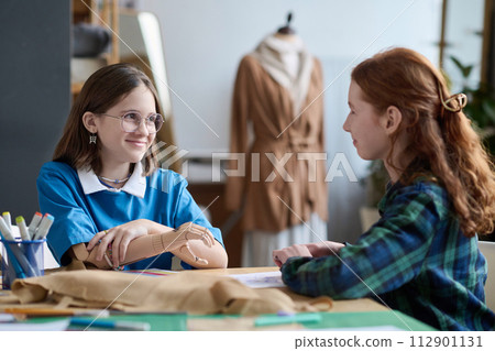 Portrait of smiling young girl with prosthetic hand chatting with friend during tailoring class at school Portrait of smiling young girl with prosthetic hand chatting with friend during tailoring class at school 112901131