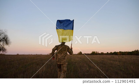 Soldier of ukrainian army running with raised blue-yellow banner on field at dusk. Young male military in uniform jogging with flag of Ukraine at meadow. Victory against russian aggression concept 112901491