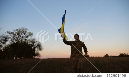 Soldier of ukrainian army running with raised blue-yellow banner on field at dusk. Young male military in uniform jogging with flag of Ukraine at meadow. Victory against russian aggression concept Soldier of ukrainian army running with raised blue-yellow banner on field at dusk. Young male military in uniform jogging with flag of Ukraine at meadow. Victory against russian aggression concept 112901496