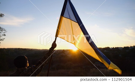 Male soldier stands with raised flag of Ukraine against background of beautiful sunset. Male ukrainian army soldier with a lifted blue-yellow banner in honor of the victory against russian aggression 112901504