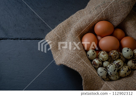 Fresh chicken and quail eggs in a basket on a sack, wooden table.Selective focus 112902312
