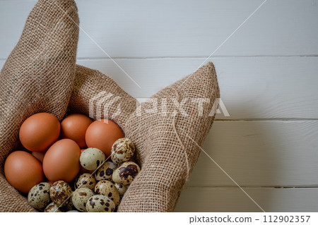Fresh chicken and quail eggs in a basket on a sack, wooden table.Selective focus 112902357