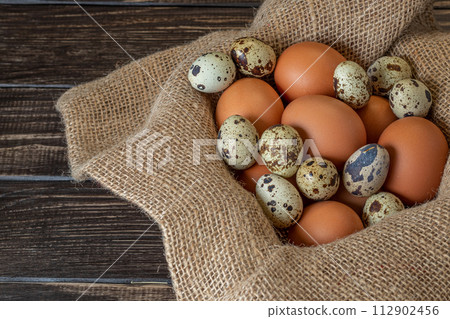 Fresh chicken and quail eggs in a basket on a sack, wooden table.Selective focus 112902456