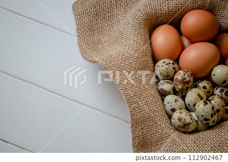 Fresh chicken and quail eggs in a basket on a sack, wooden table.Selective focus 112902467