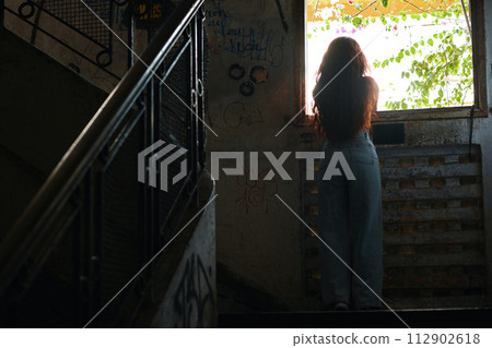 Young woman with long hair looking though window of abandoned and decaying building 112902618