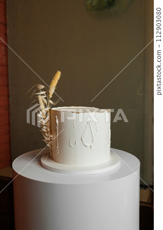 Festive one tiered birthday white cake in rustic style, decorated dry wheat spikelets and vanilla cream icing on stand at reception restaurant. Wedding, anniversary party. Catering service in event Festive one tiered birthday white cake in rustic style, decorated dry wheat spikelets and vanilla cream icing on stand at reception restaurant. Wedding, anniversary party. Catering service in event 112903080