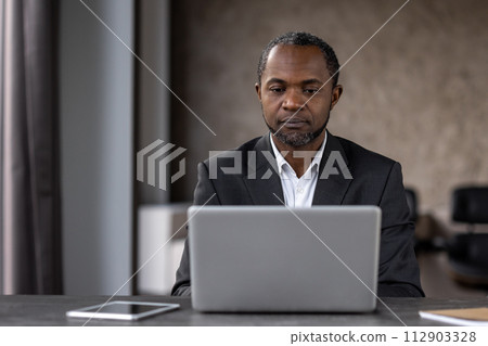 Serious focused male businessman working inside office with laptop, mature experienced african american man typing on keyboard, thinking reading from monitor. 112903328