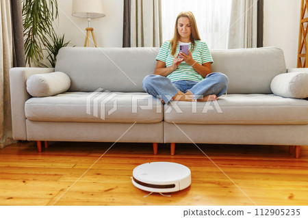 Woman using smartphone on couch with robotic vacuum cleaner on the floor Woman using smartphone on couch with robotic vacuum cleaner on the floor 112905235